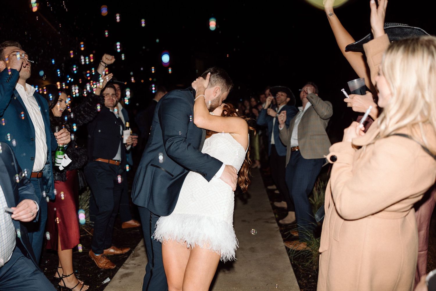 A crowd of people dance and celebrate at a nighttime wedding reception with colorful lighting and decor.