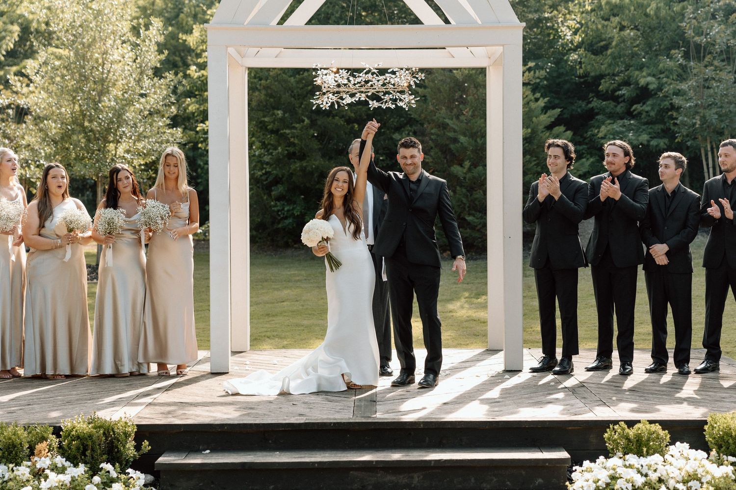 Wedding party members stand in formal attire under a white outdoor gazebo during a sunny garden ceremony.