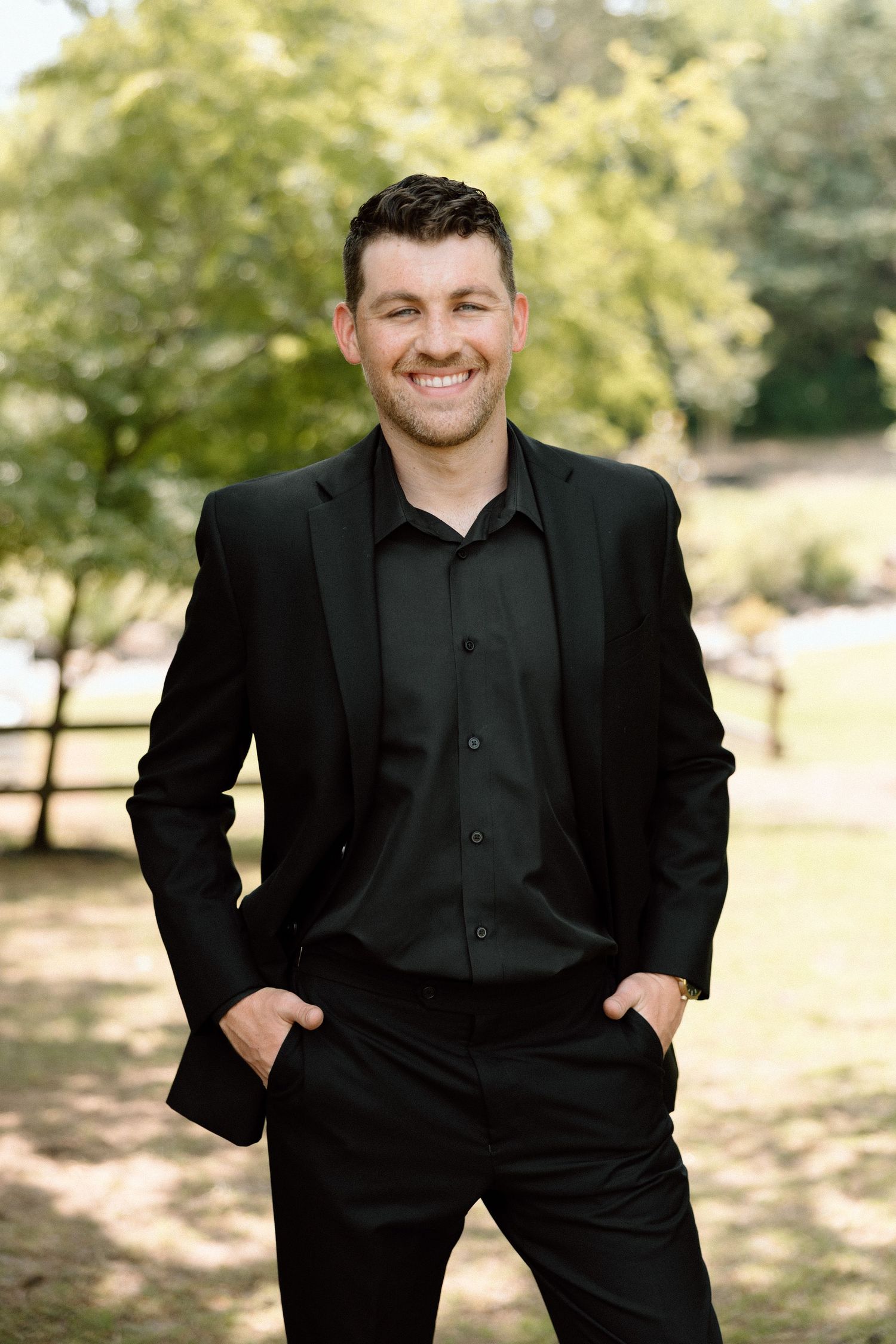 Professional portrait series showing a person in a black suit and dress shirt posing casually outdoors in a park setting.
