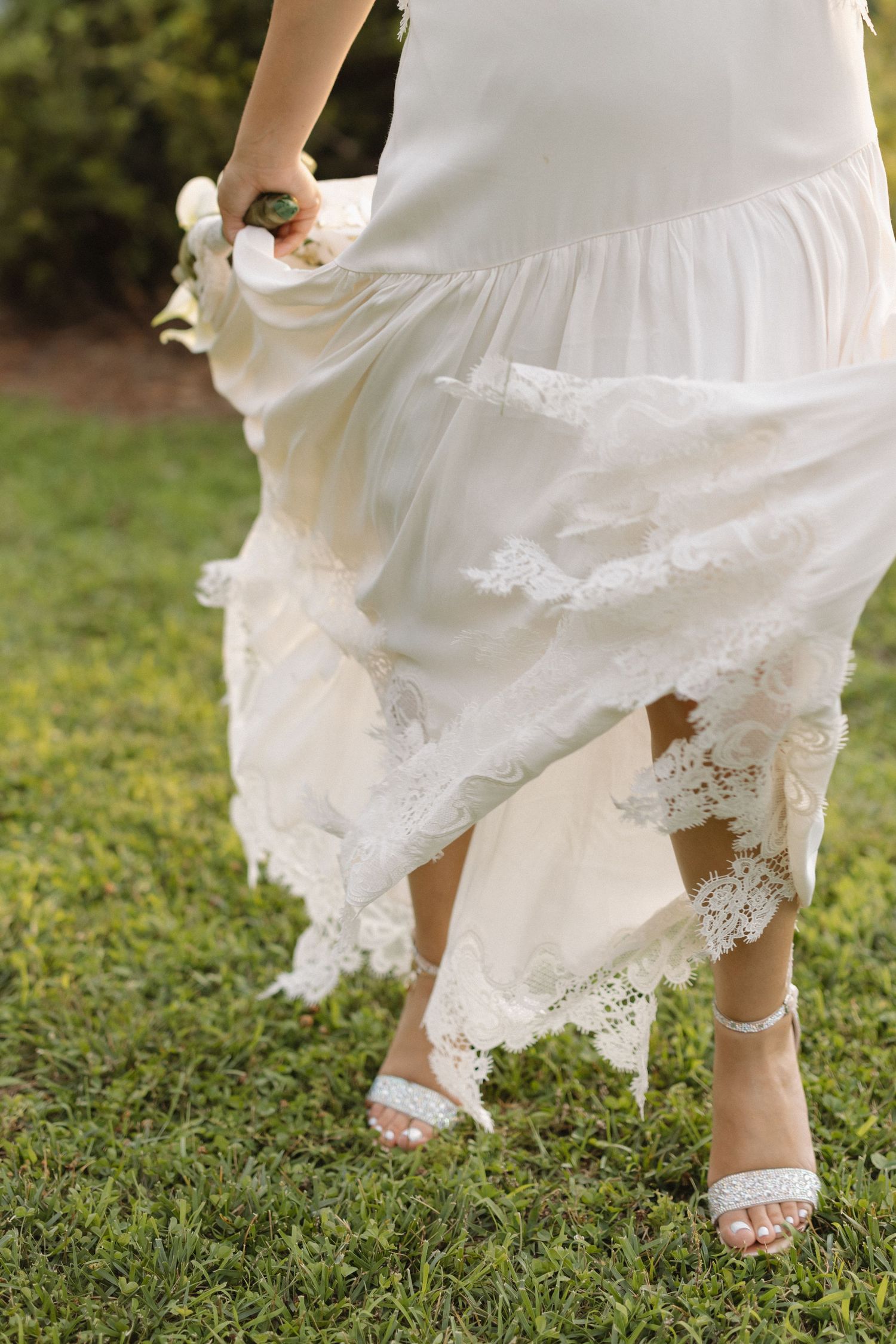 Close-up of a bride's lace wedding dress hem and embellished white heels on grass.