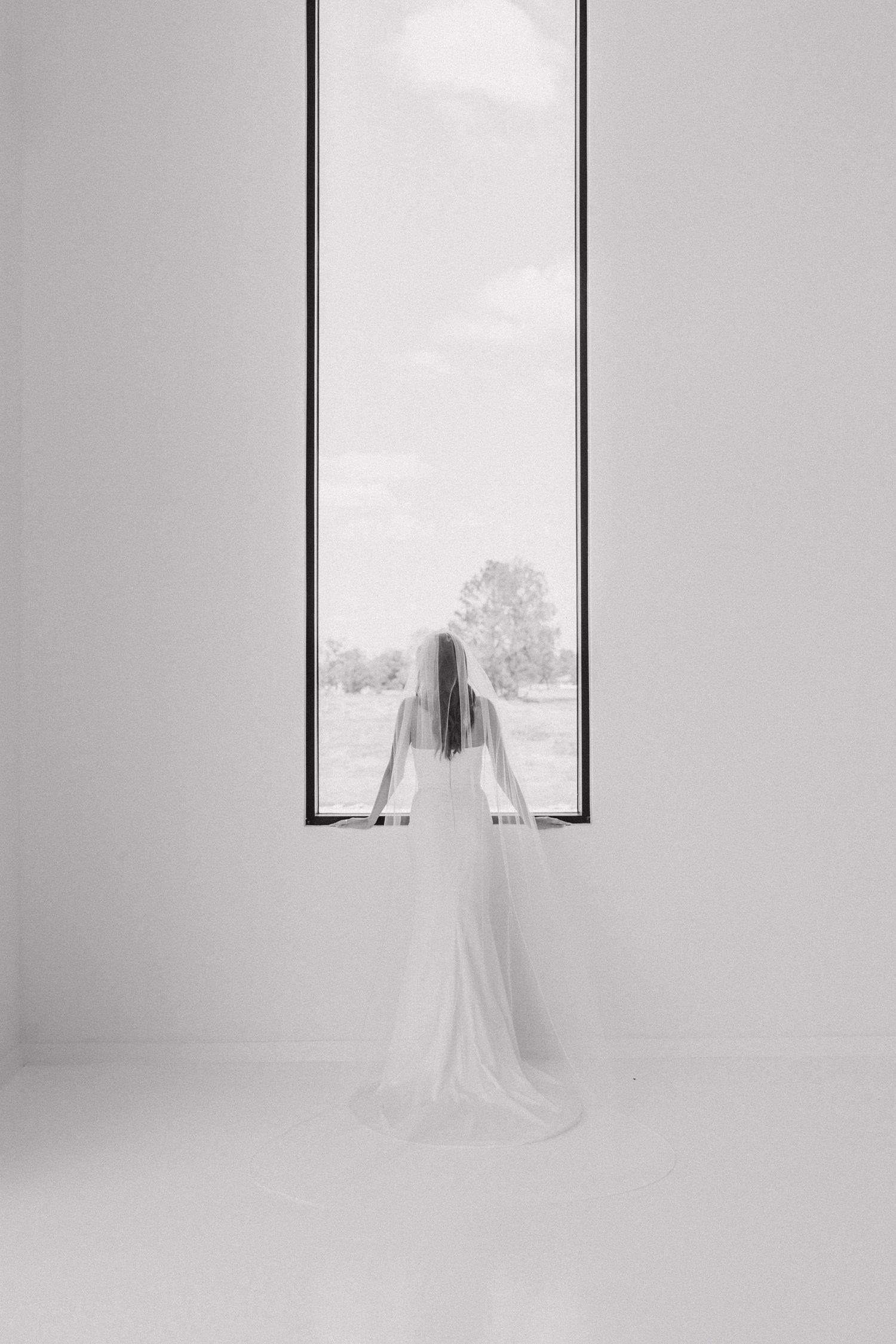 Artistic black and white photo of a bride in flowing veil standing at a minimalist window.
