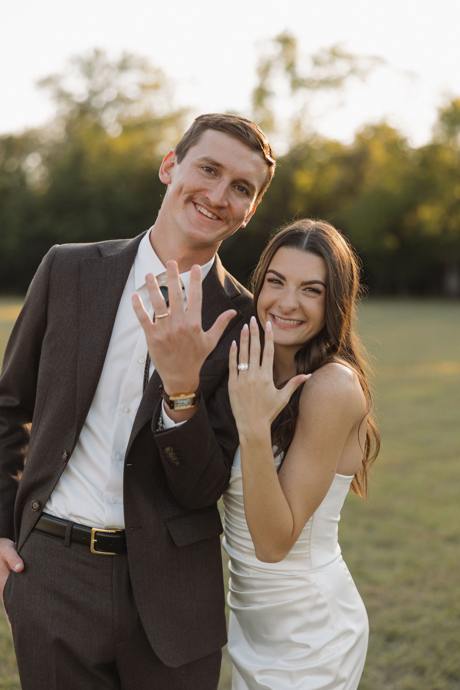 Newlywed couple showing off their rings with joyful expressions in an outdoor setting.