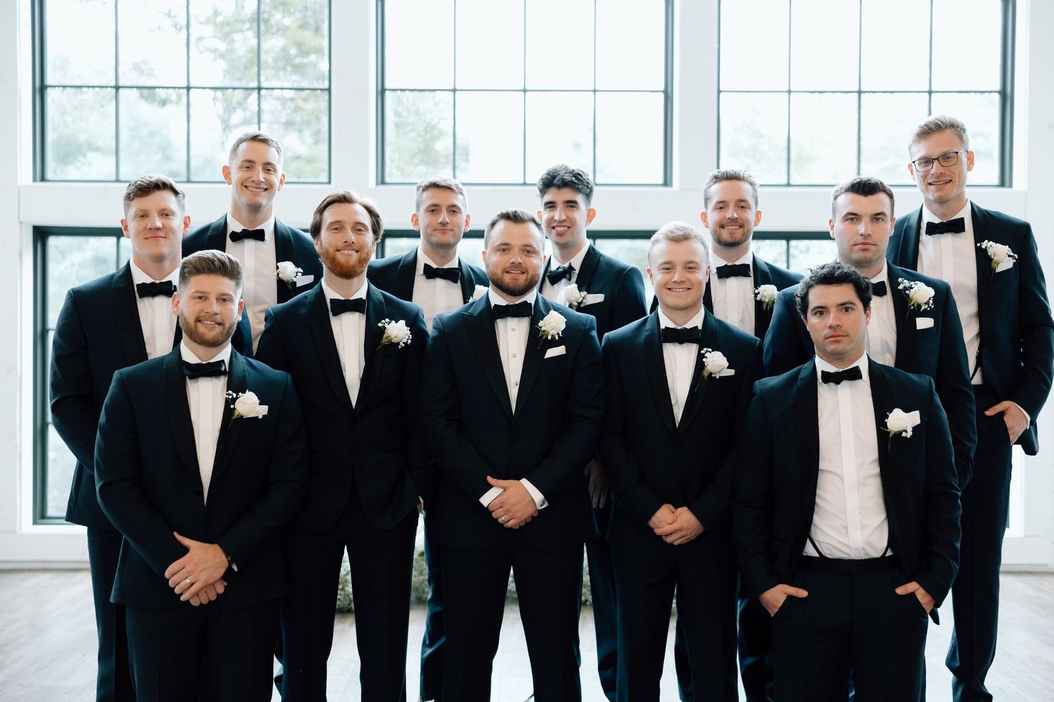 A group of groomsmen in black tuxedos and bow ties stand in a line at a formal wedding venue.
