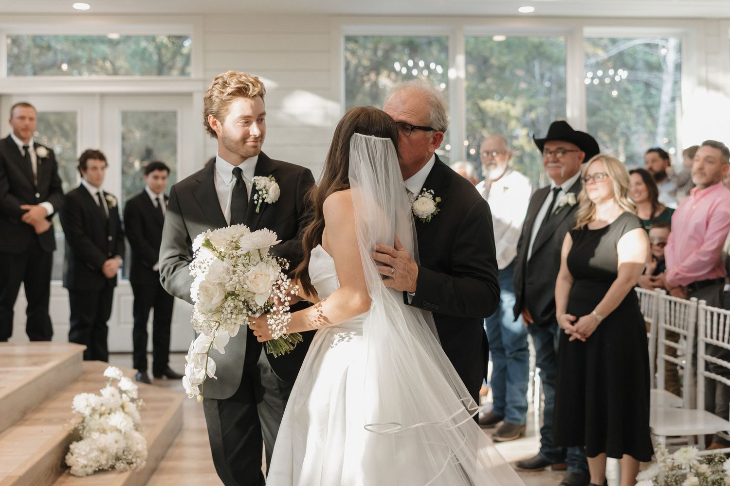 Wedding ceremony guests seated in white chairs watch as the couple exchanges vows in an elegant indoor venue.