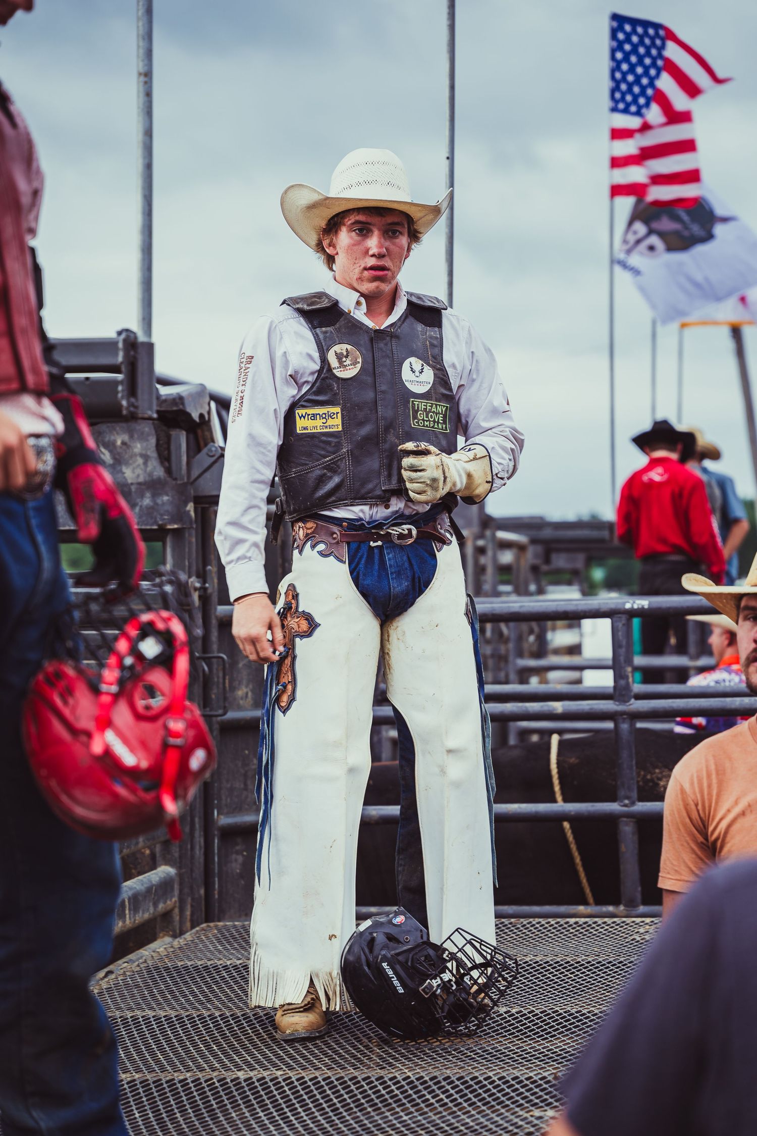 Young rodeo cowboy in white chaps, vest, and hat stands at chutes with American flag backdrop.