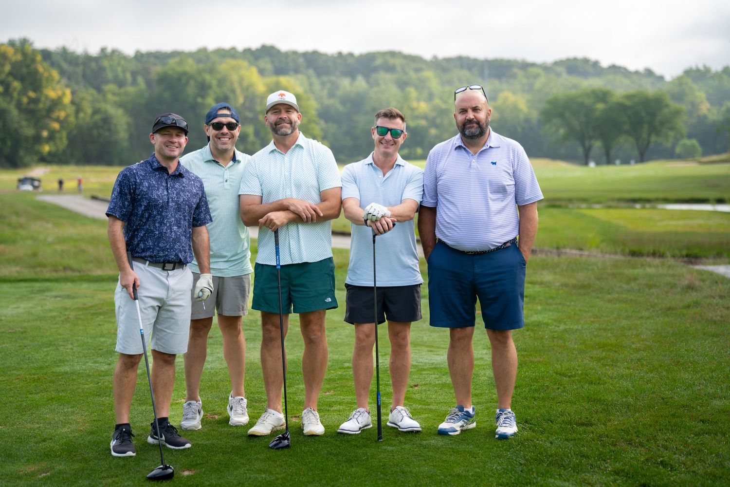 Five men in golf attire smiling together on a lush green golf course on a sunny day.