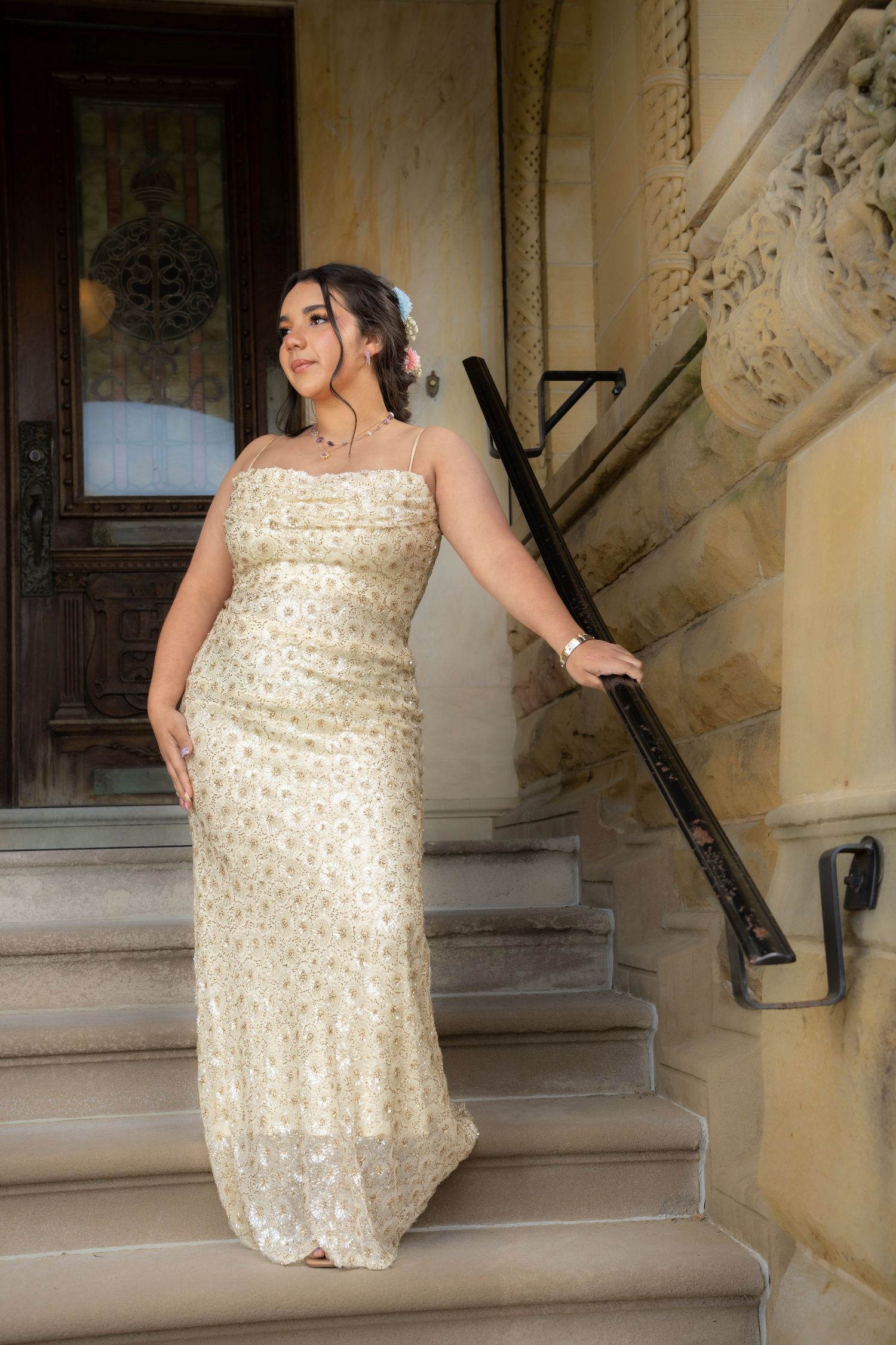 Young woman in elegant gold sequin gown poses on stone steps beside ornate wooden door.