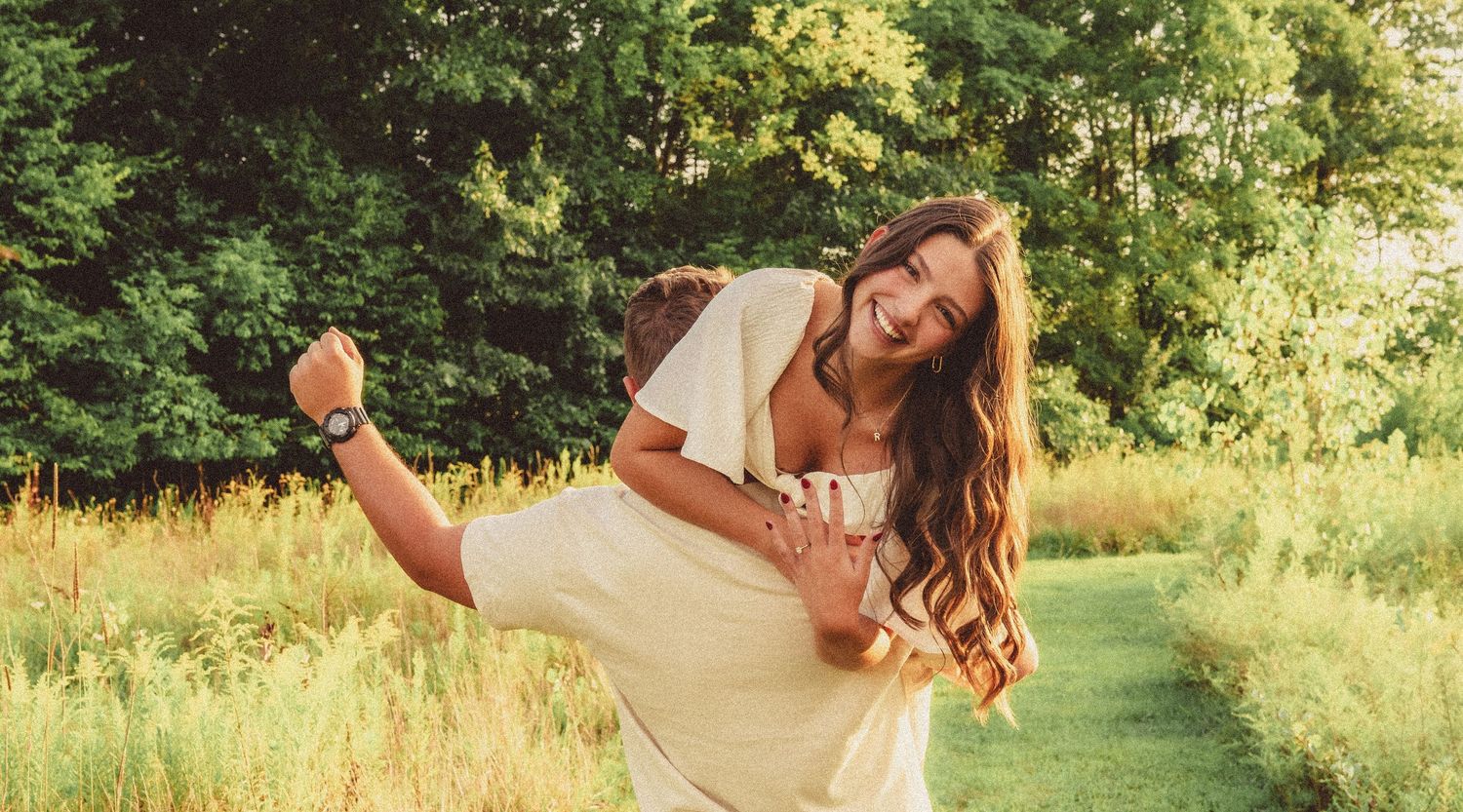 Joyful man lifting his smiling fiancee in a green meadow surrounded by trees.