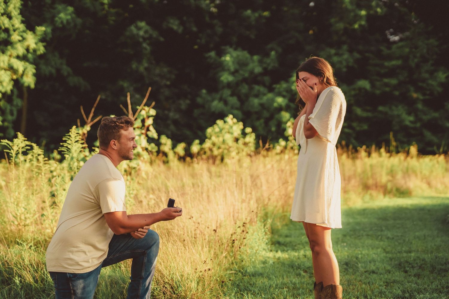 Man presenting engagement ring while kneeling before surprised woman in white dress outdoors.