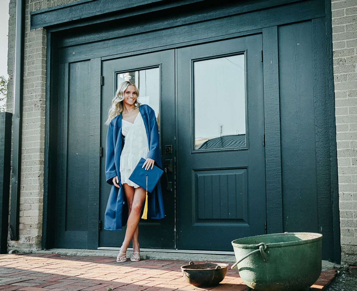 Blonde woman in white dress holding blue graduation gown beside dark double doors.