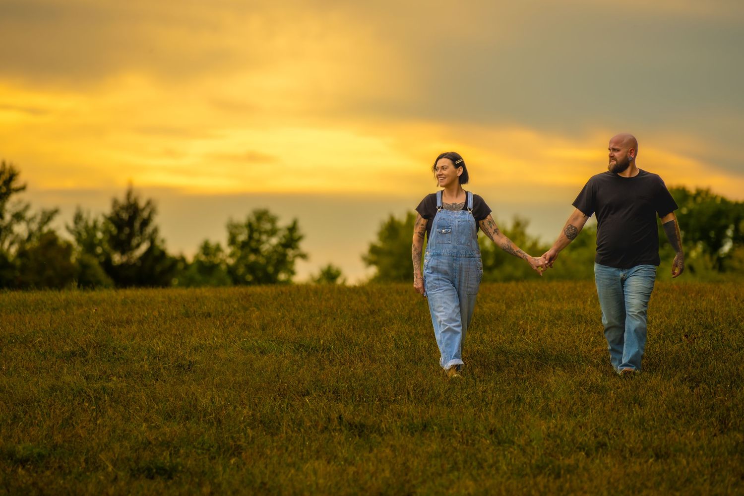 Couple holding hands walking across a green field beneath a vivid orange sunset sky.