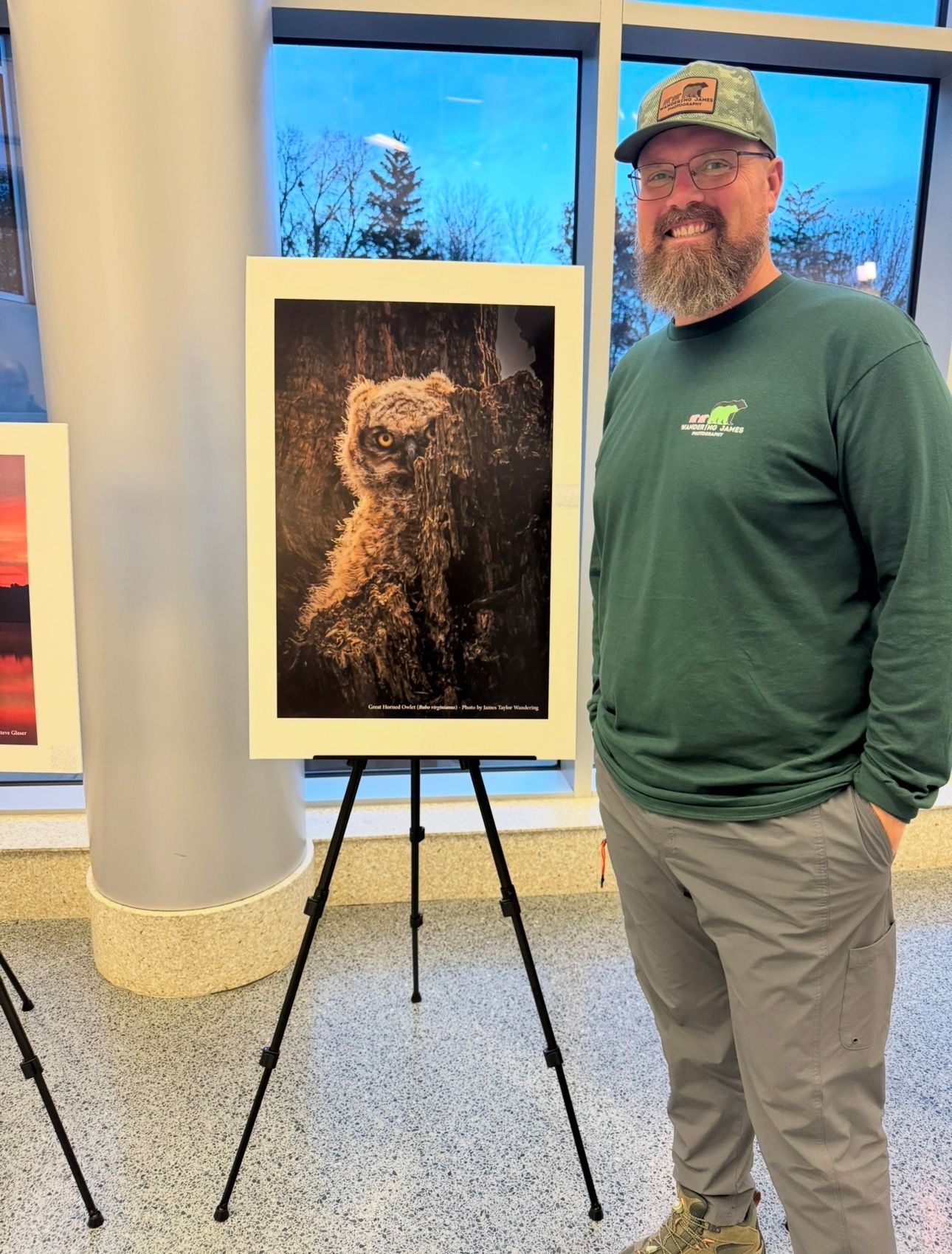 Bearded man in green shirt and cap standing beside a framed owl photo on an easel in an indoor gallery.