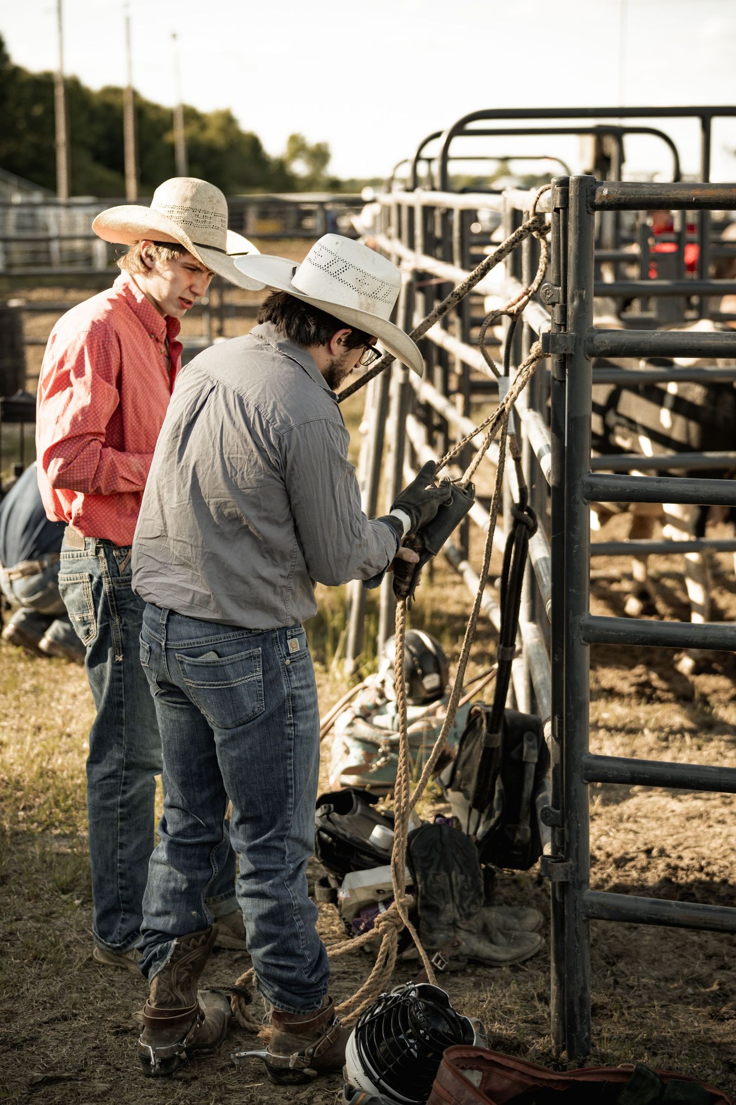 Two cowboys in white hats prepare rodeo equipment at metal livestock pens before a rodeo event.