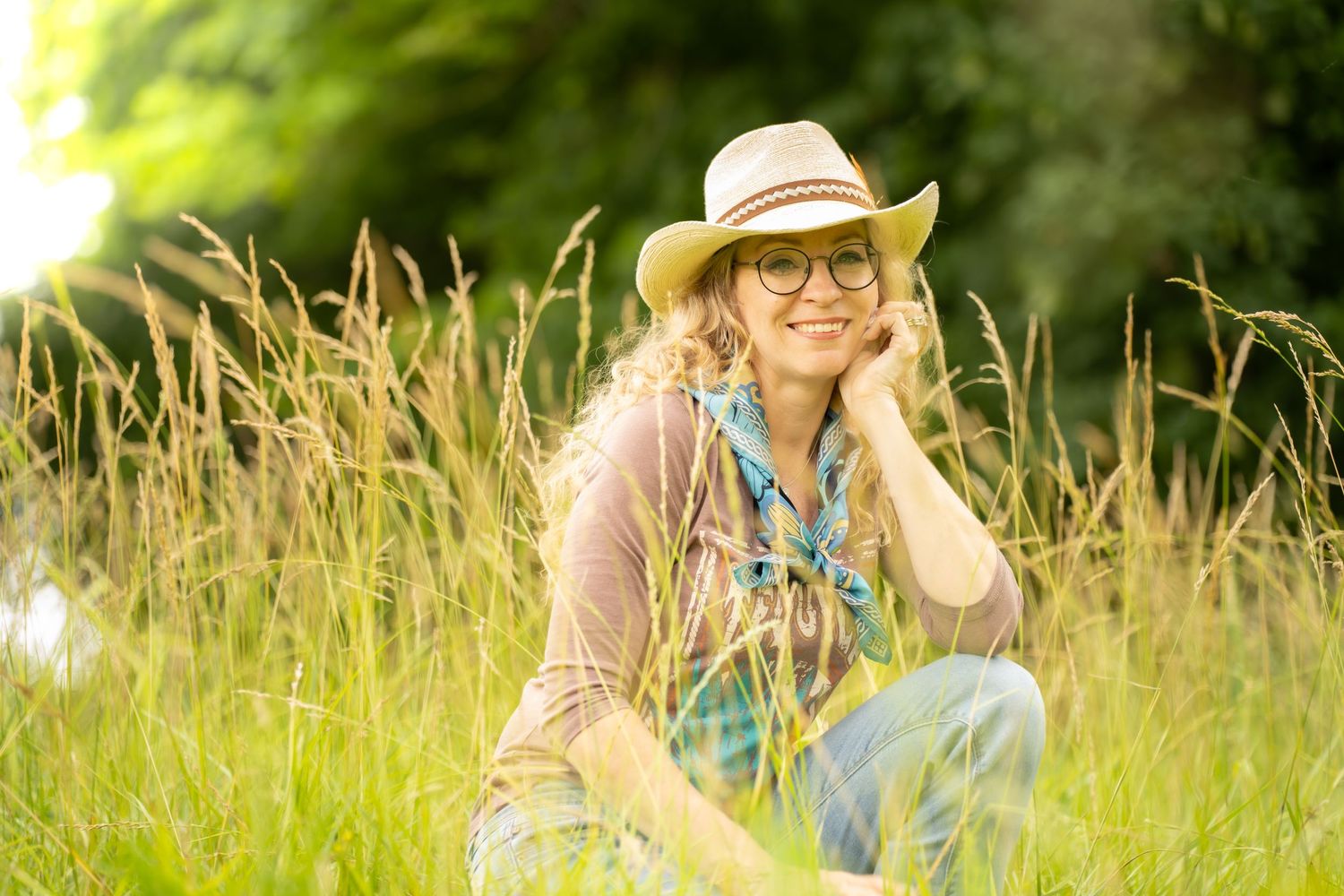 A smiling woman in a straw hat and glasses sits in tall green grass on a sunny day.