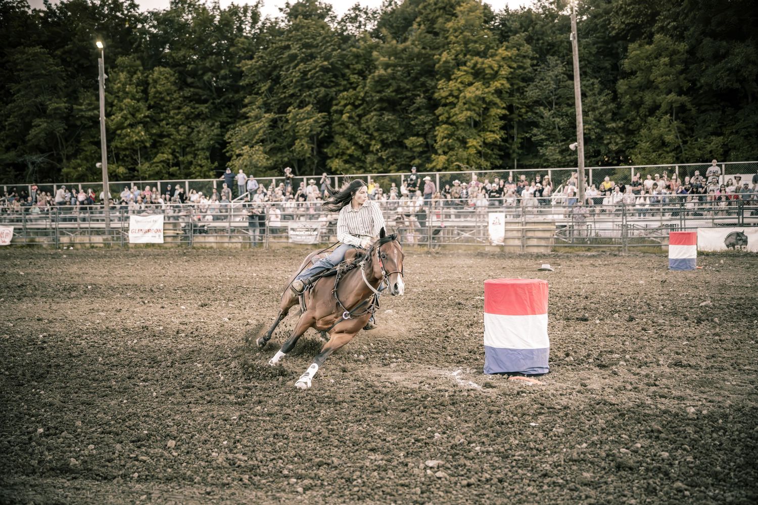 Barrel racer on a brown horse rounds a red-white-blue barrel in a dirt arena.