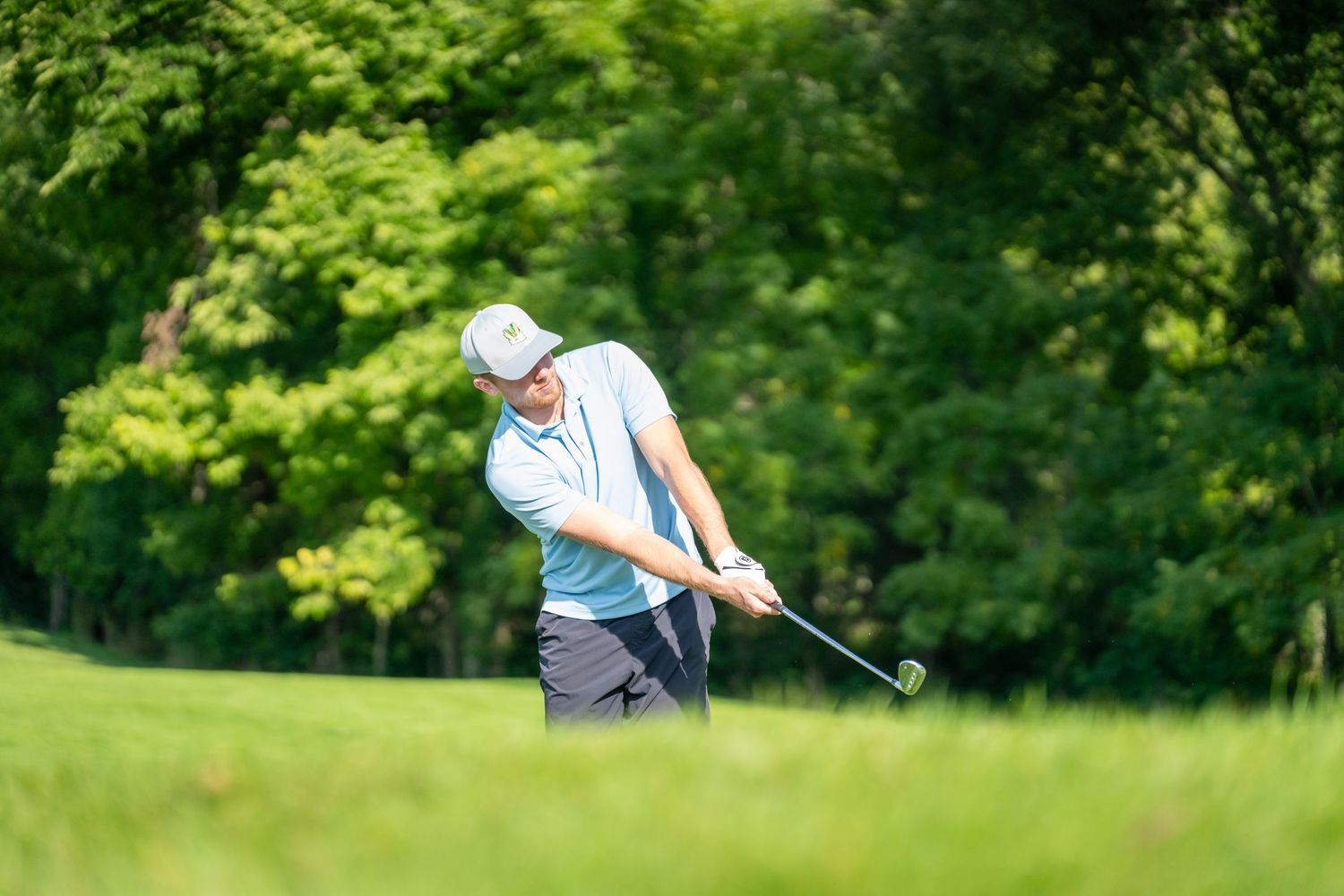 Golfer mid-swing on a lush green golf course surrounded by trees on a sunny day.