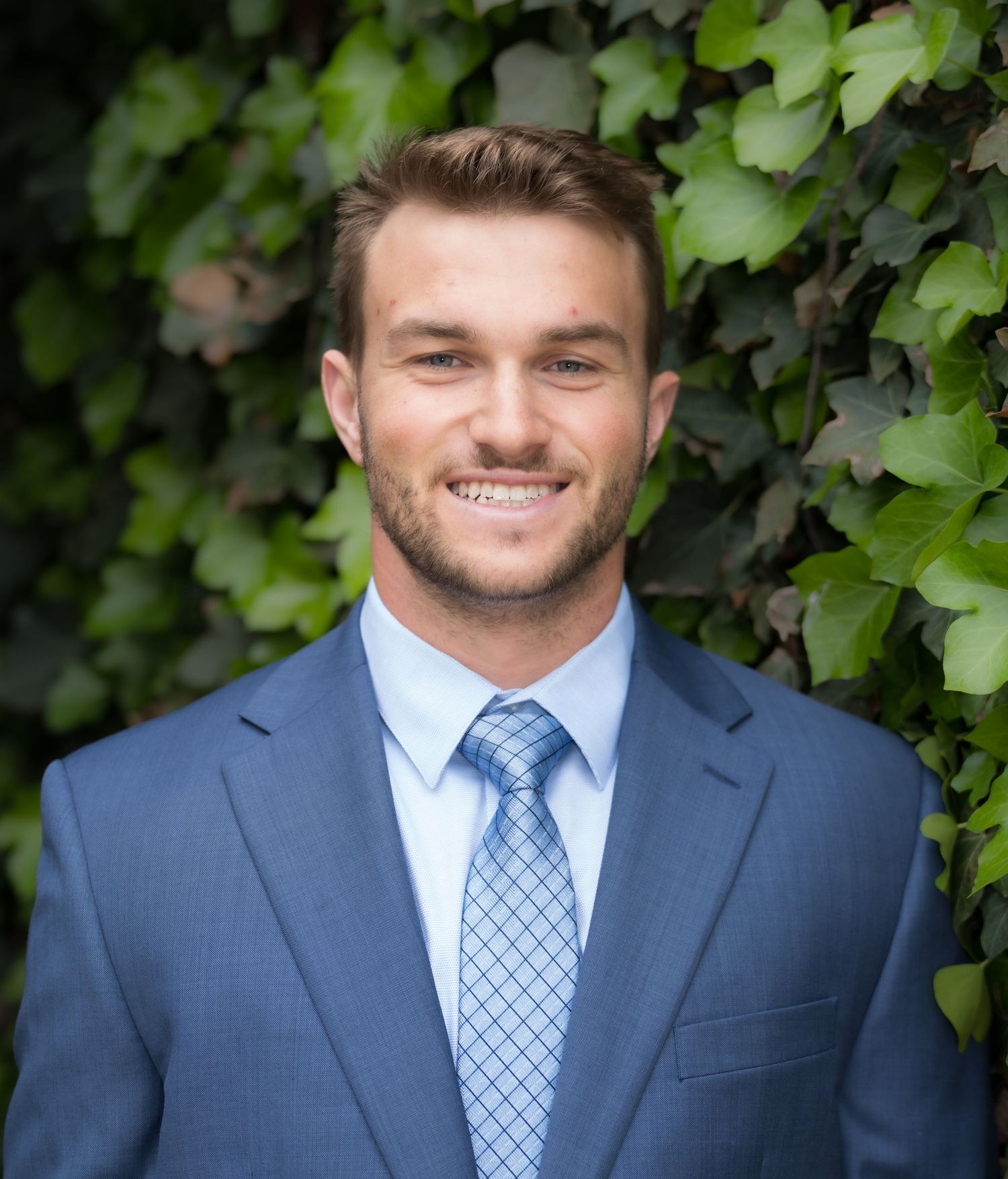Young professional man in blue suit and tie smiling outdoors with green ivy background.