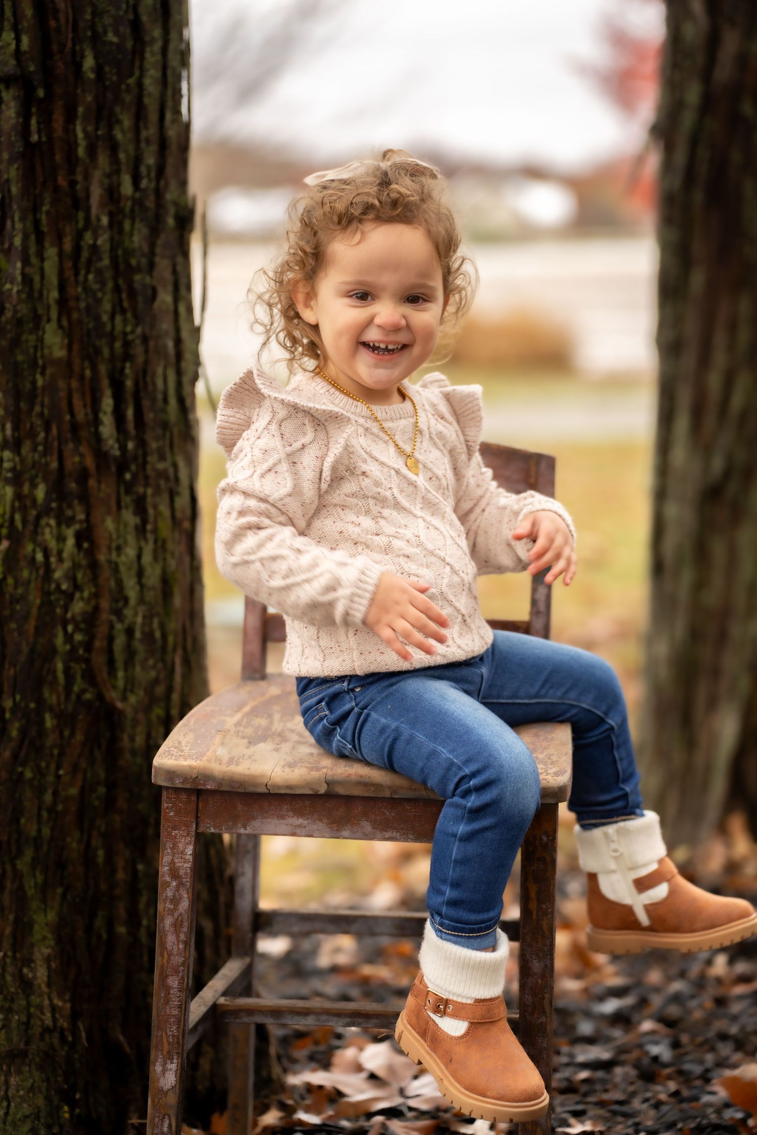 Adorable curly-haired toddler girl smiling while sitting on a rustic wooden chair outdoors.