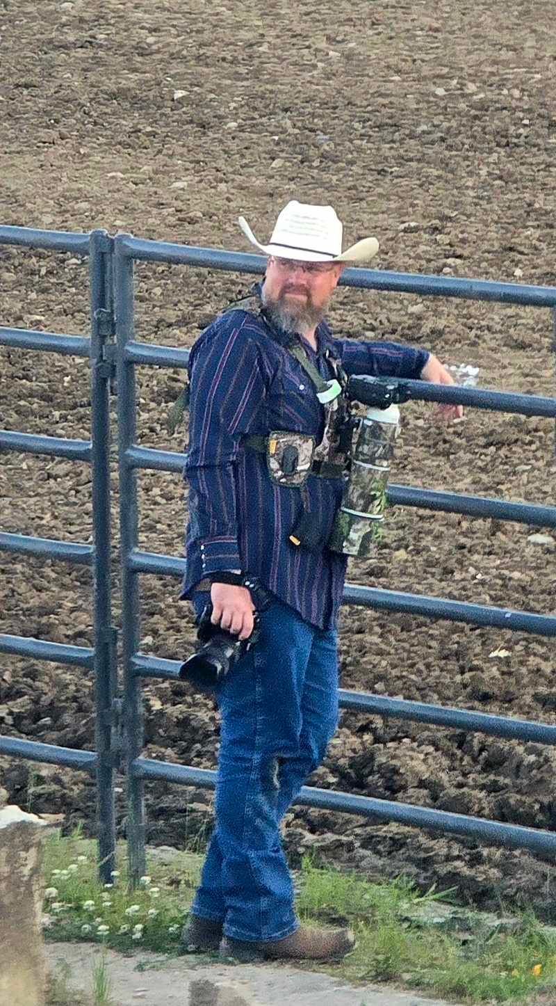 Bearded man in white cowboy hat, blue shirt, jeans, and boots leaning on metal fence at rodeo with cameras.