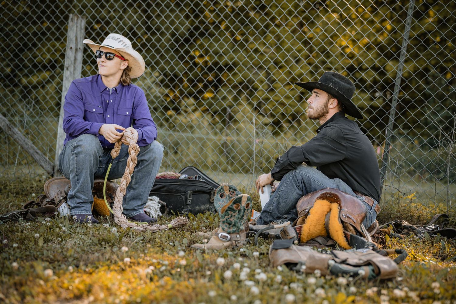 Two cowboys in hats sit and chat against a chain-link fence at an outdoor rodeo event.