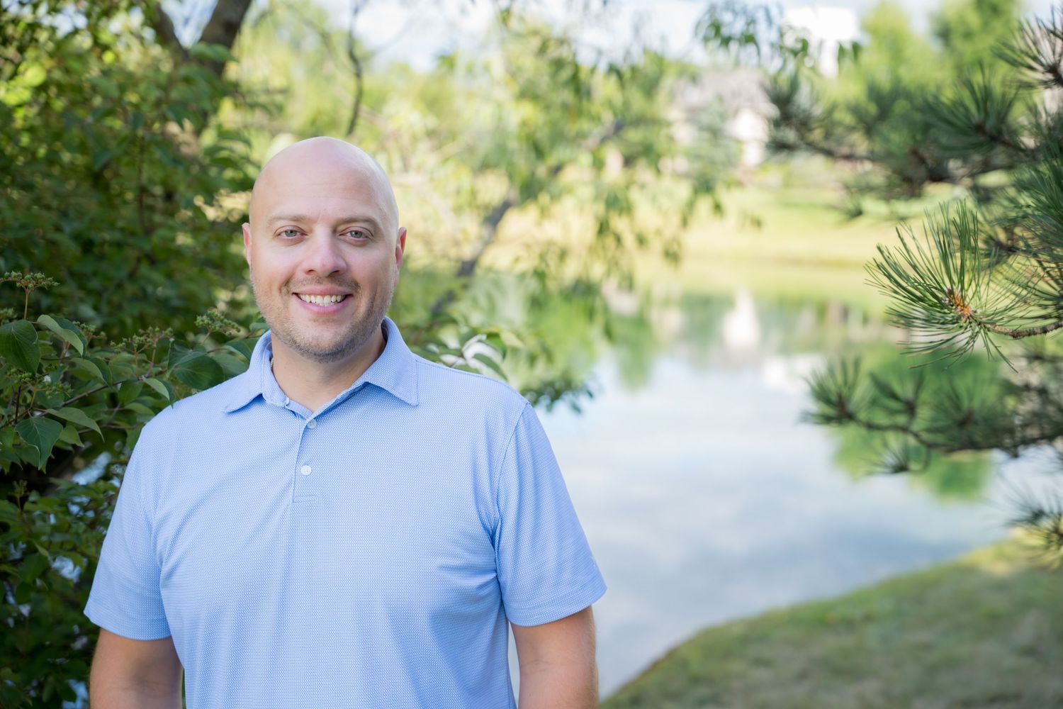 Smiling bald man in light blue polo shirt standing outdoors near a serene pond surrounded by lush green trees.