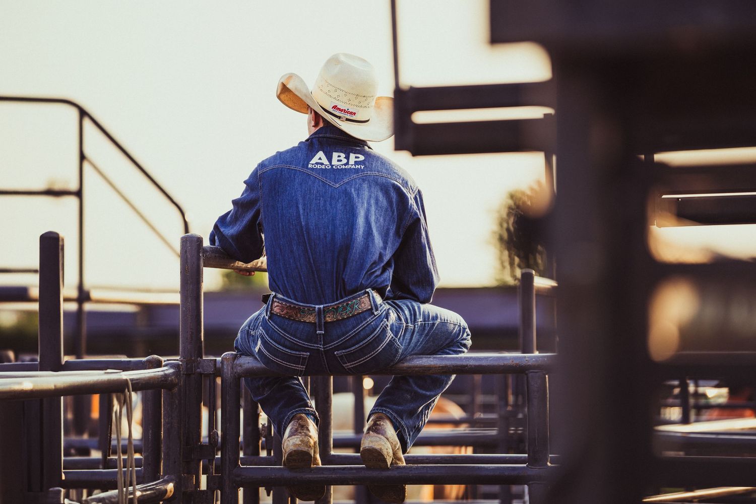 Cowboy in denim jacket with ABP logo sits on metal fence wearing white hat at a rodeo arena at sunset.