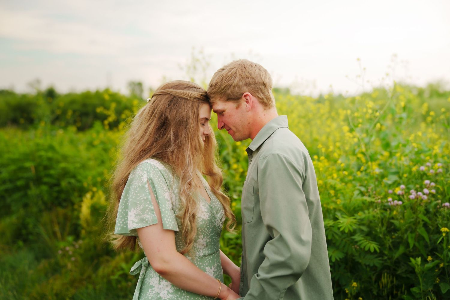 Romantic couple in sage green outfits standing face to face in a lush wildflower meadow during golden hour.