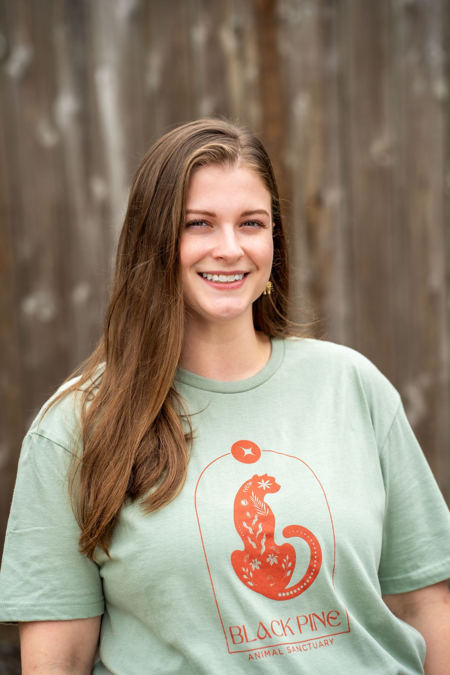 Smiling young woman wearing a mint green Black Pine Animal Sanctuary t-shirt standing outdoors.