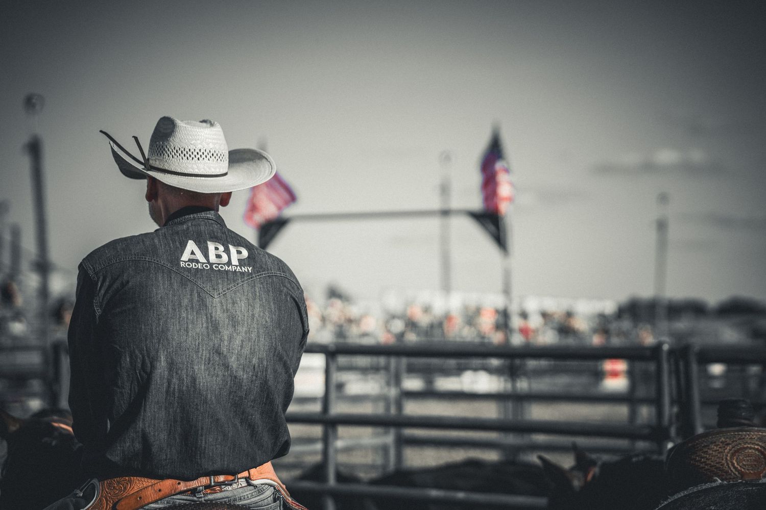 Cowboy in ABP jacket and white hat sits on horseback at a rodeo with American flag in background.