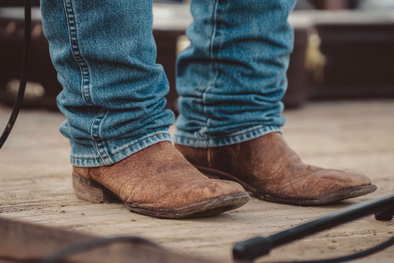 Worn brown leather cowboy boots with blue jeans on a wooden floor.