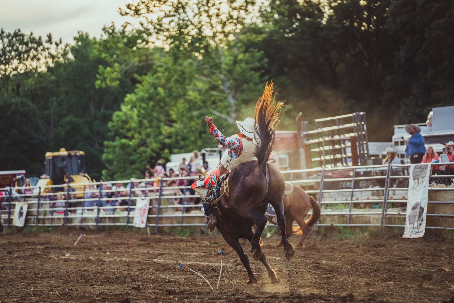 A cowboy rides a bucking bronco at a rodeo competition with spectators watching from behind the fence.