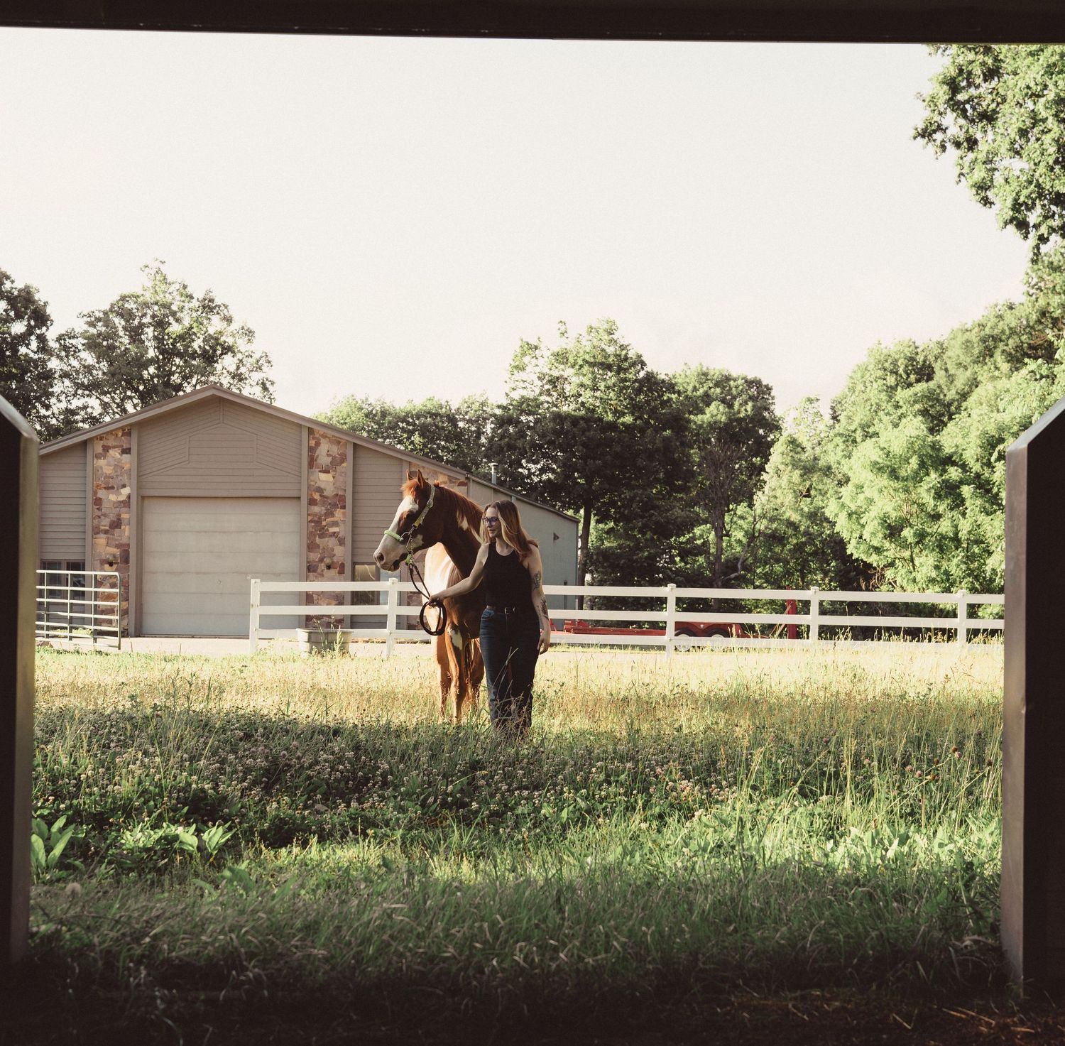 A person stands with a brown horse in a lush green pasture beside a barn and white fence on a farm.