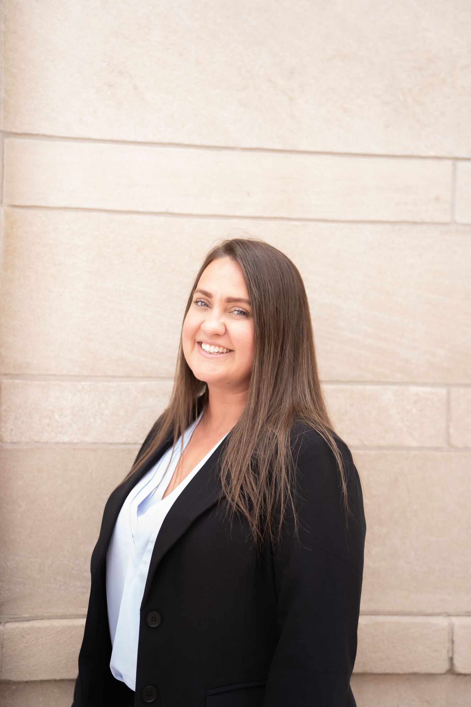 Professional woman in black blazer and white blouse smiling confidently against a stone wall background.