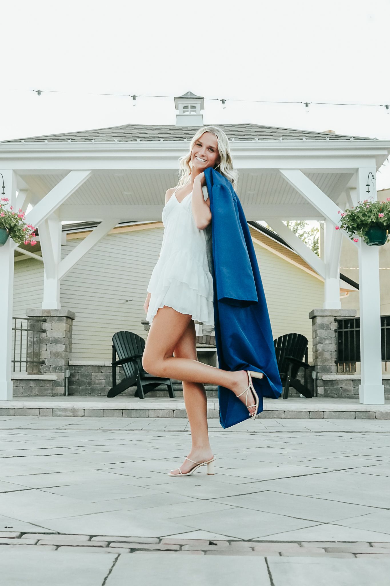 Smiling blonde woman in white dress twirling blue graduation gown near outdoor pavilion.