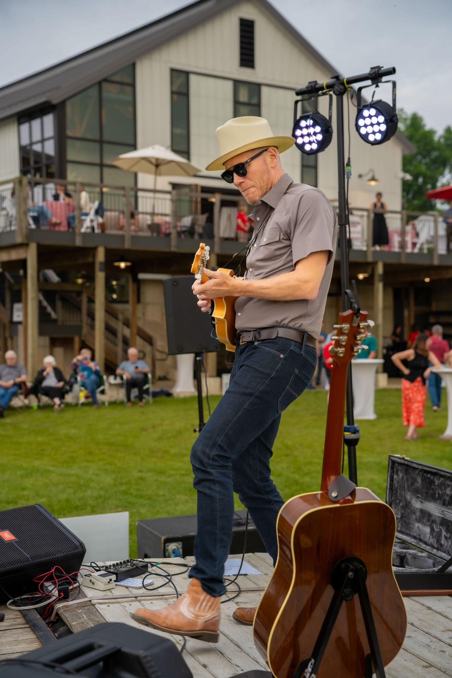 Outdoor guitarist in straw hat and sunglasses performs on stage with acoustic guitar leaning nearby at a backyard event.