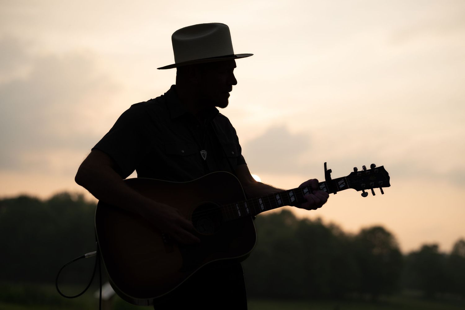 Silhouette of a guitarist wearing a wide-brim hat playing acoustic guitar against a dramatic sunset sky.