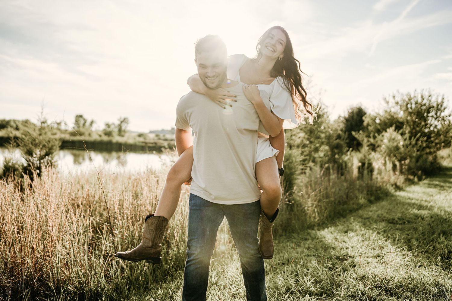 Man giving woman a piggyback ride near a lake in golden sunlight among tall grass.