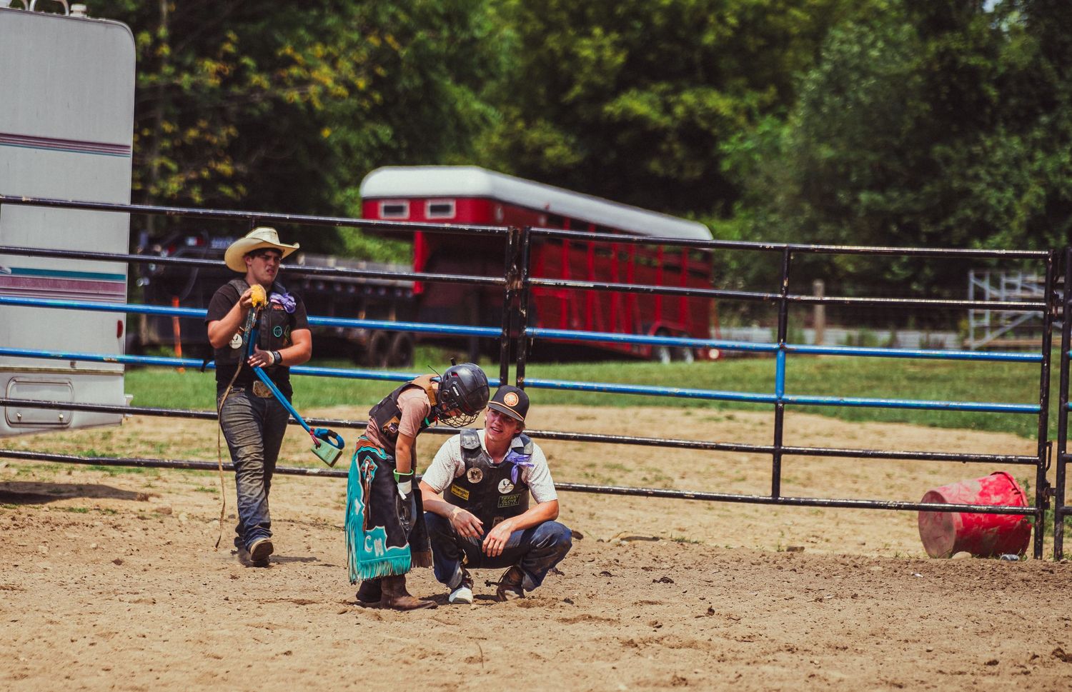 Two rodeo participants crouch in a sandy arena near a fence, with trees and a red trailer visible in the background.