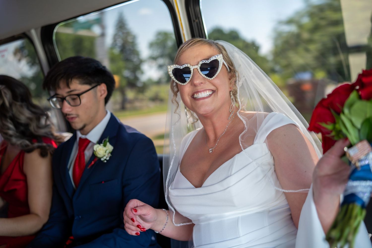 Smiling bride wearing heart-shaped white sunglasses and veil holds red bouquet while sitting in a wedding car.