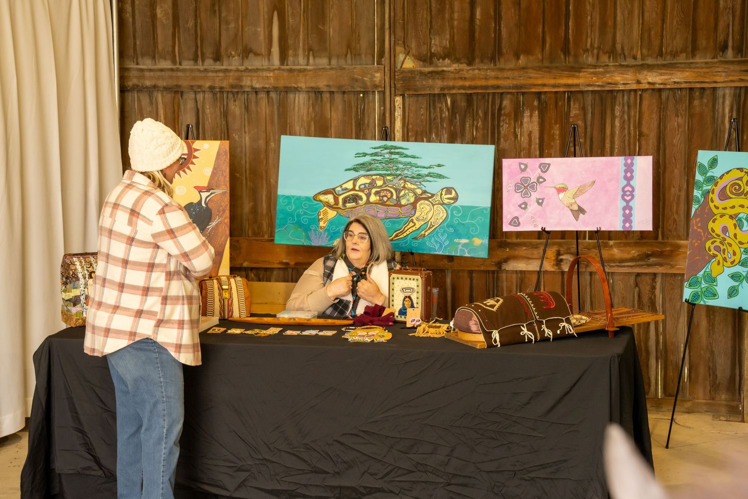 Artist sits behind a table displaying colorful paintings and handmade goods inside a rustic wooden barn.