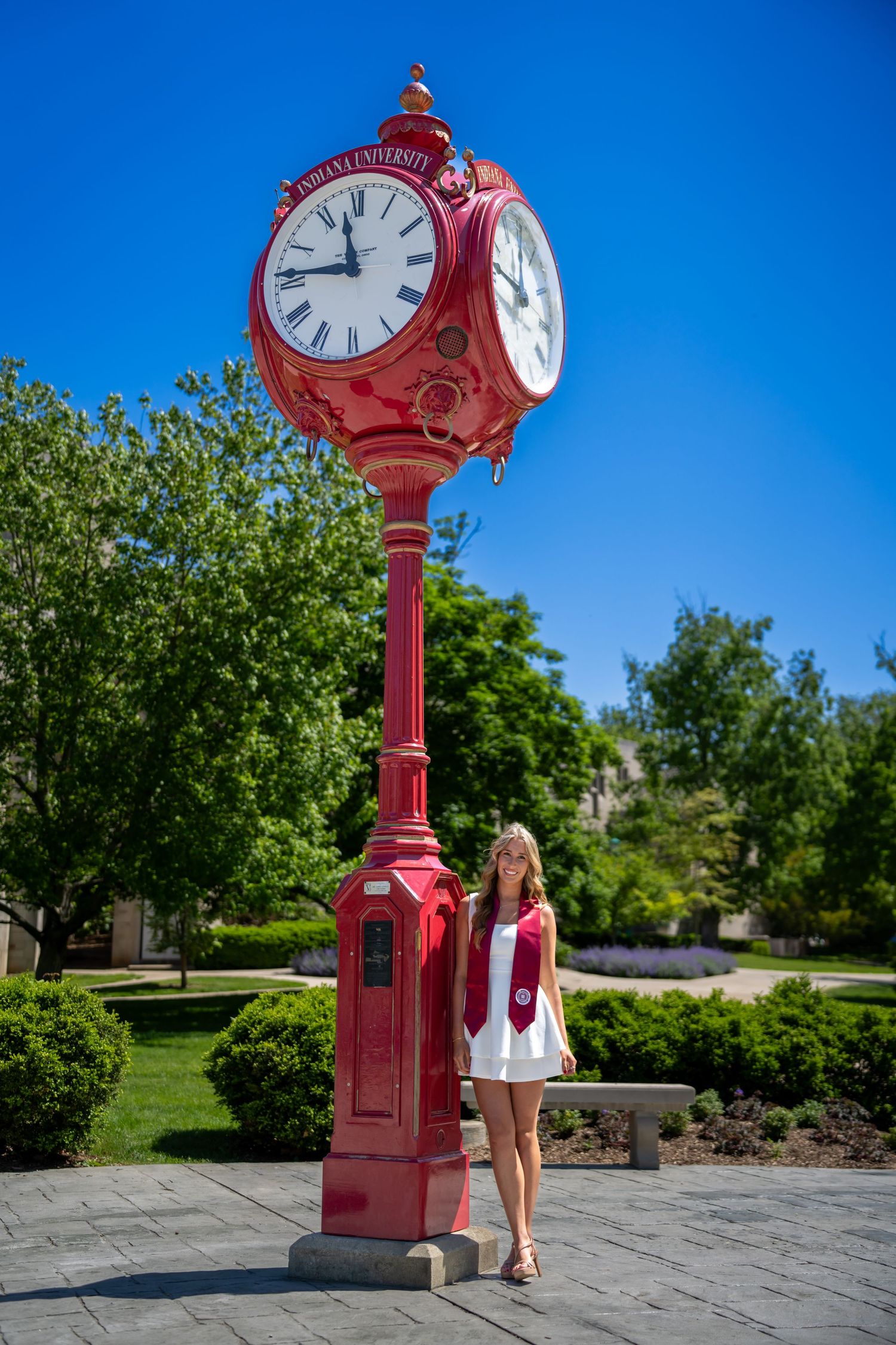 Blonde graduate in white dress standing beside iconic red Indiana University campus clock.