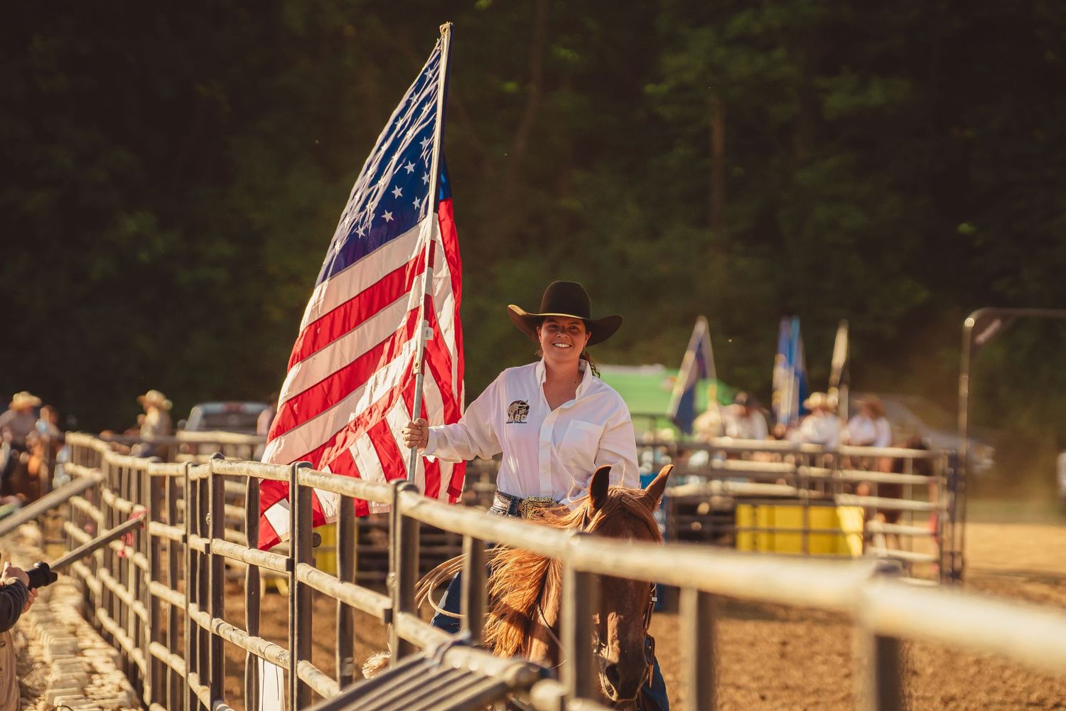 Smiling cowgirl on horseback carries a large American flag through a rodeo arena at golden hour.