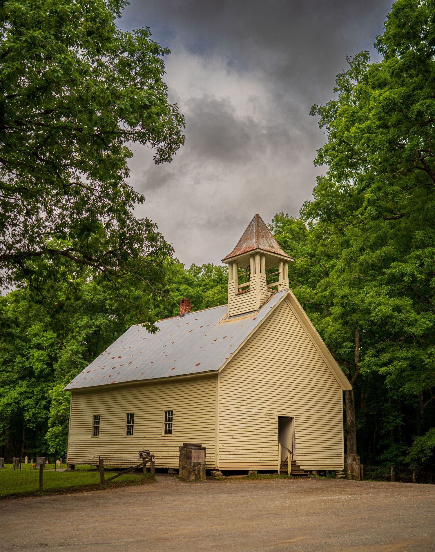 Historic white wooden church with bell tower surrounded by lush green trees under dramatic cloudy skies.