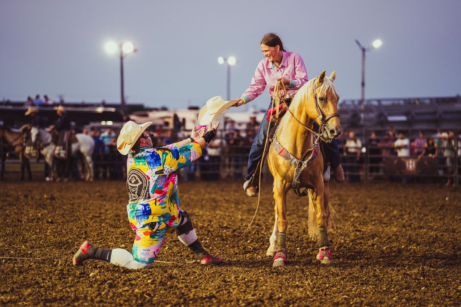 A colorful clown kneels before a rider on a palomino horse at a lively rodeo event under bright arena lights.