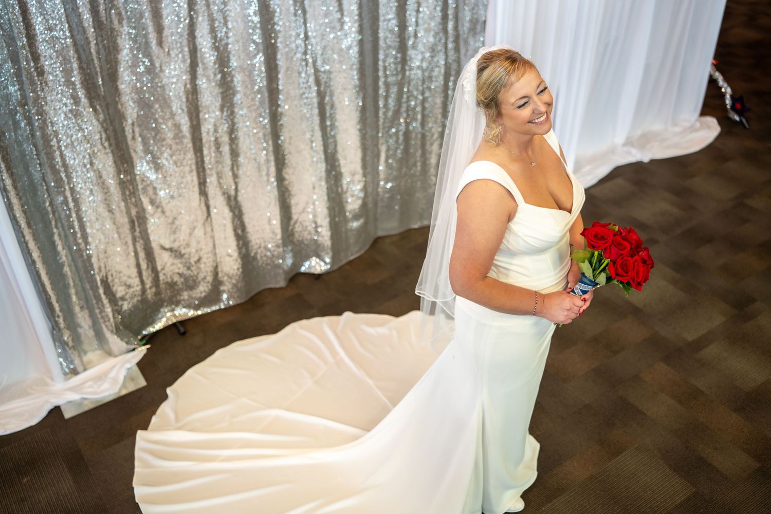 Smiling bride in white gown holding red bouquet stands near silver sequin backdrop on her wedding day.