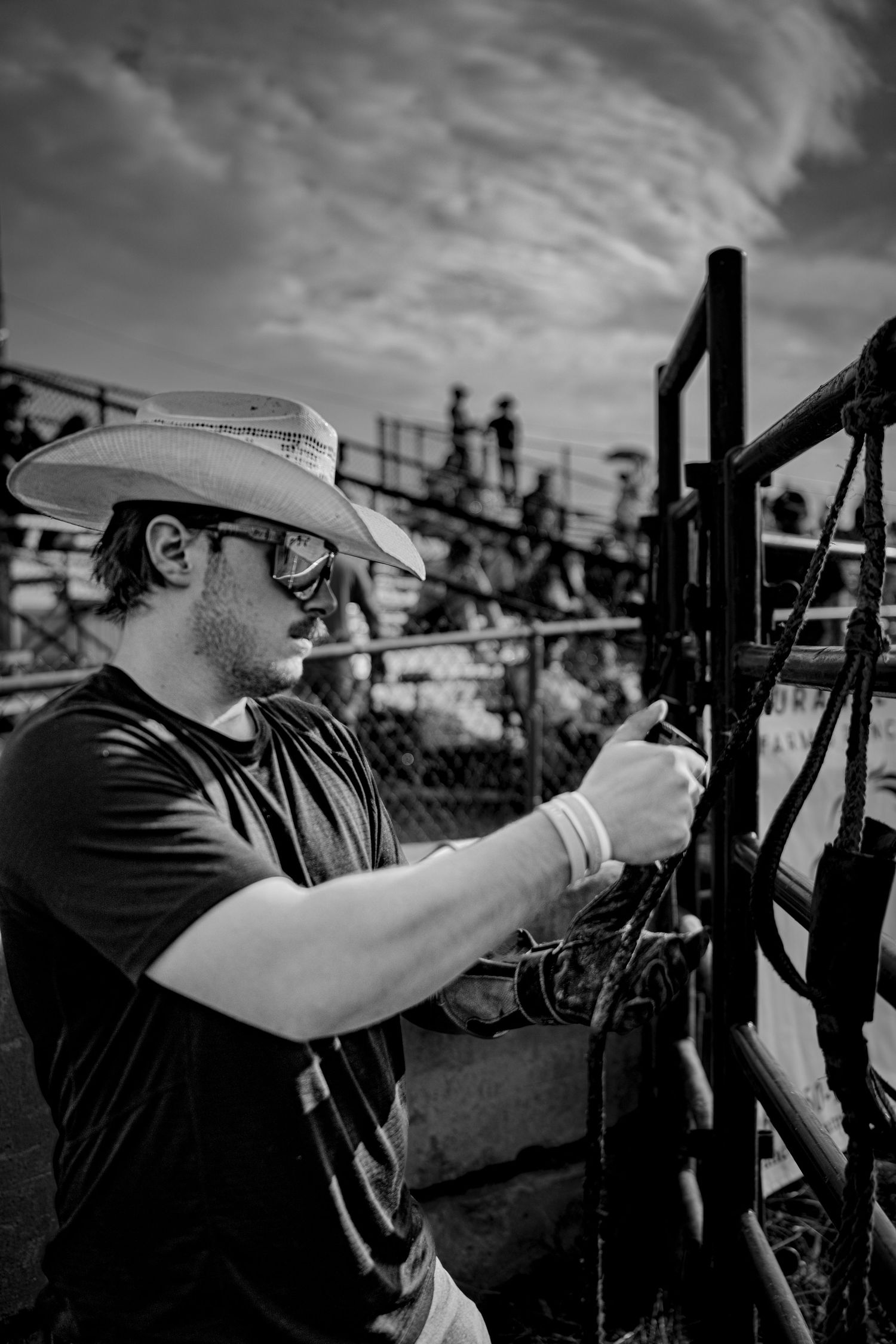 Black and white photo of a cowboy in sunglasses and hat working with ropes at a rodeo fence.