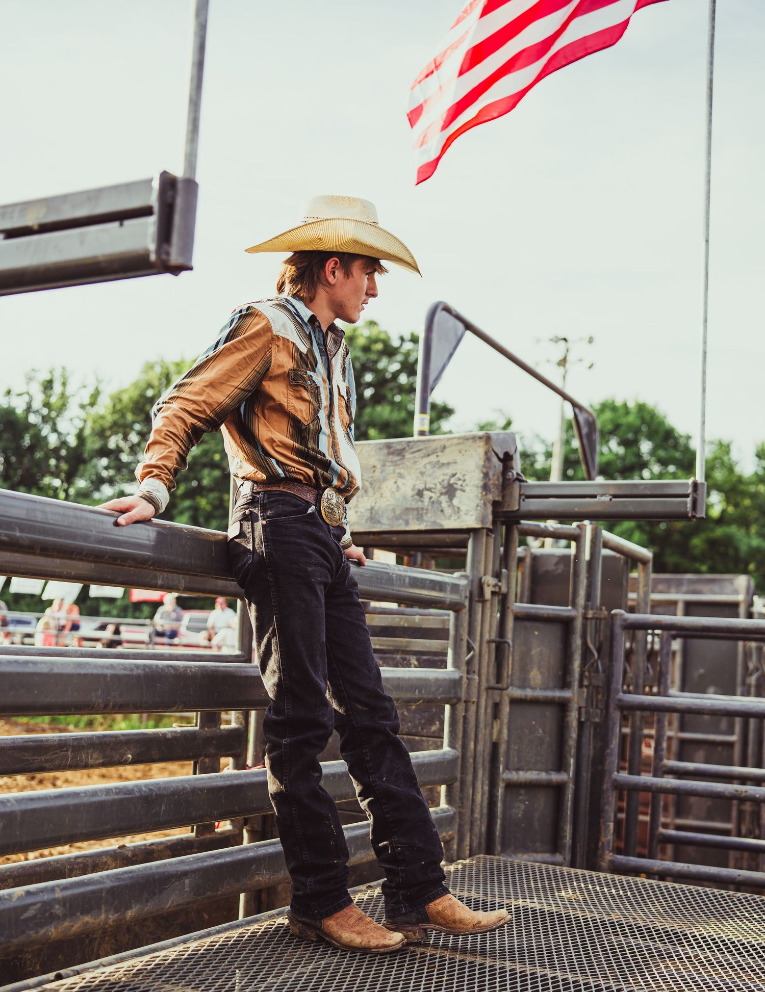 A young cowboy in a straw hat and vest stands on a rodeo fence beside an American flag.