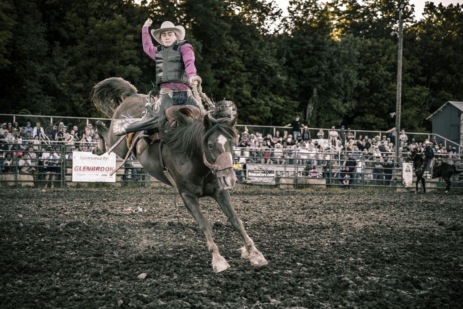 Cowboy riding a bucking bull at a rodeo competition with crowd watching in the background.