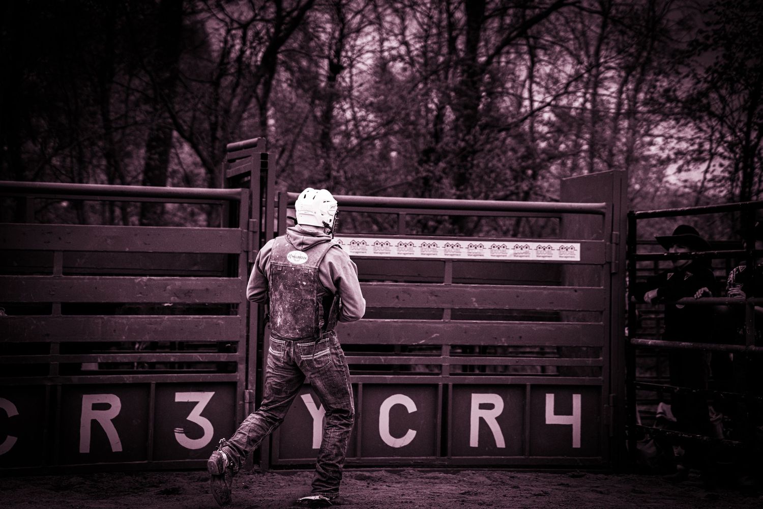 A cowboy stands before a rodeo chute gate marked CR3 and CR4 in a moody monochrome scene.