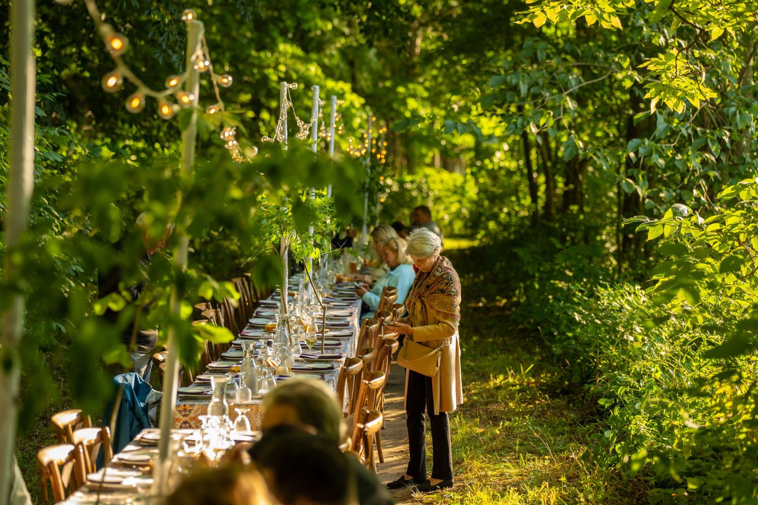 Long elegant outdoor dining table set in a lush green forest path with string lights and candles for an al fresco event.